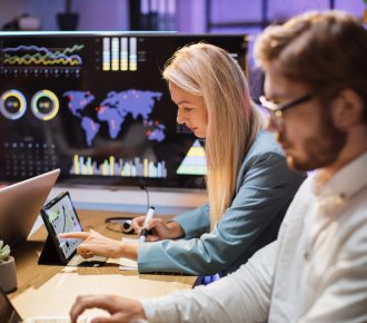 Side view of caucasian male and female financiers with modern gadgets on desk studying database of trade market together. Cooperation for common success. Evening time at office.
